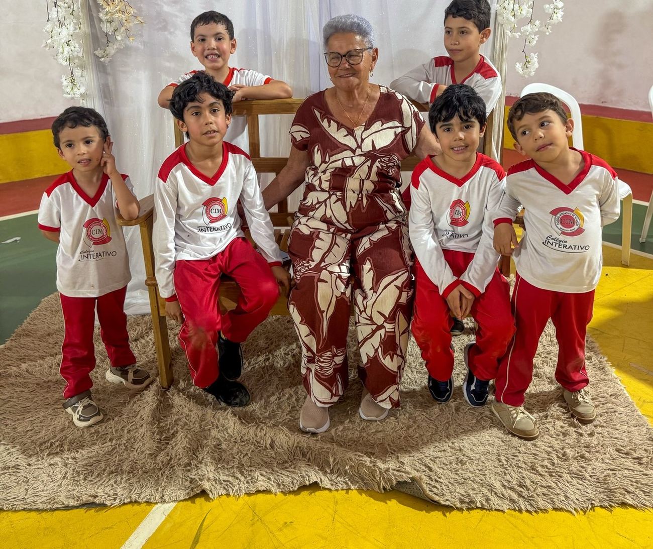 Alunos da Educação Infantil do Colégio Interativo de Maracás, de uniforme branco e vermelho, ao lado de uma avó homenageada na escola